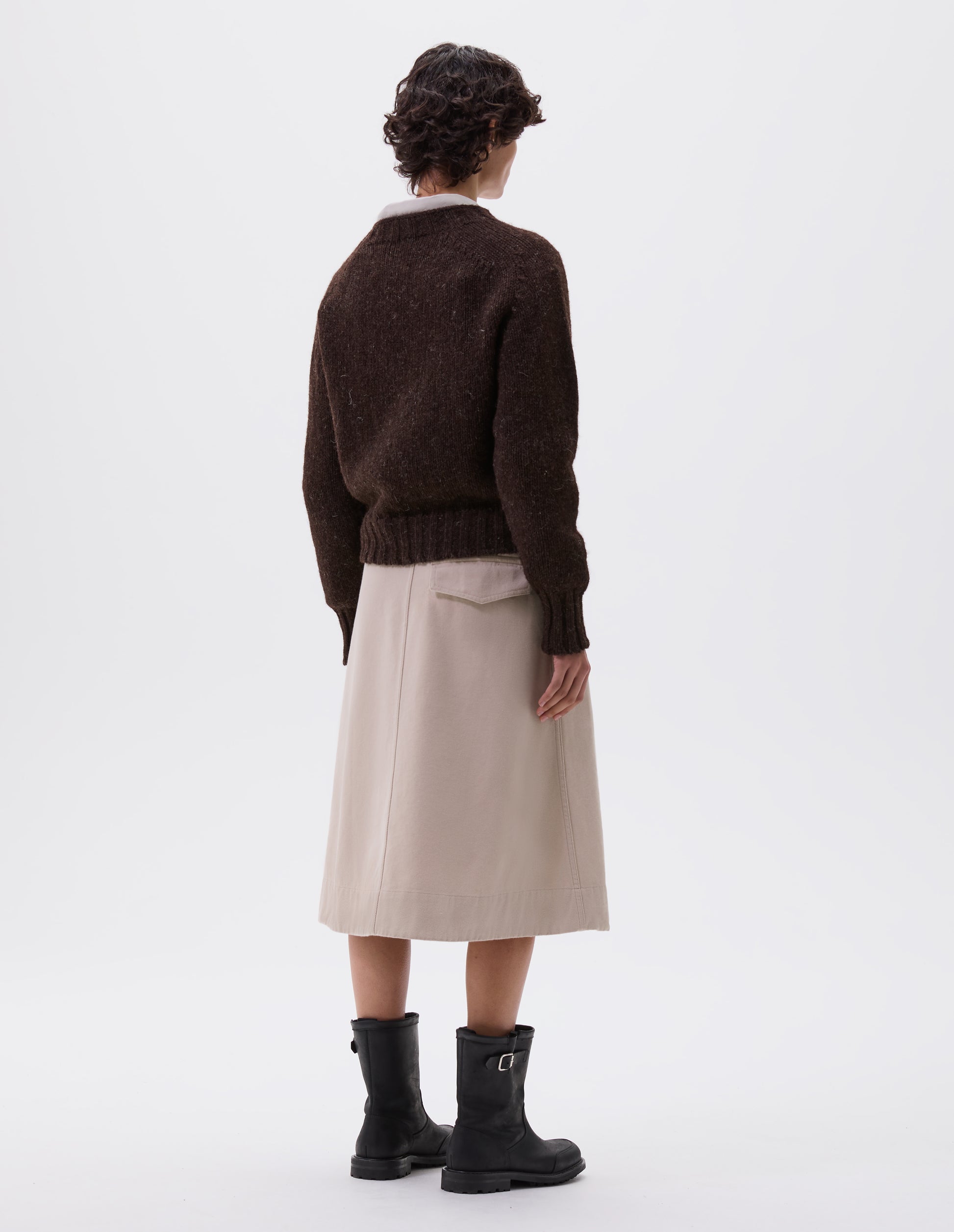 Full body image, back view of model wearing dark brown rustic wool jumper with saddle sleeve and contrasting rib on neck, cuffs and hem. Styled with beige skirt and black leather boots.