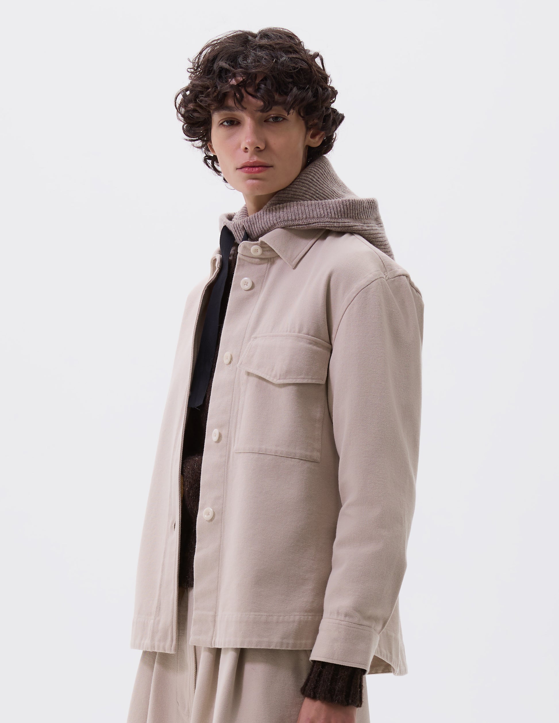 Cropped body image, front view of female model wearing beige knitted wool hood with contrasting black herringbone tape to adjust and contrast rib on face. Styled with beige jacket and matching skirt.