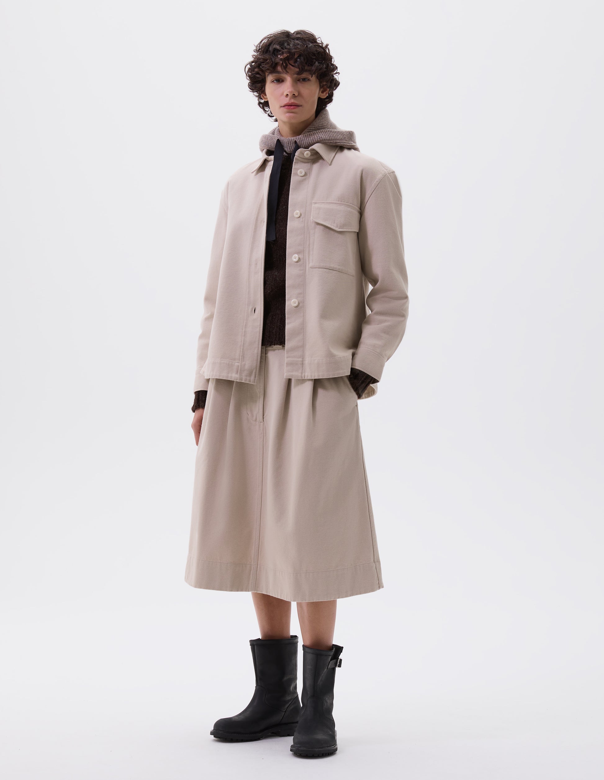 Full body image, front view of model wearing beige oversized shirt with stitch through raised placket, corozo button fastening, patch chest pocket and deep stitch hem. Styled with matching skirt, brown jumper, beige hooded scarf and black leather boots.