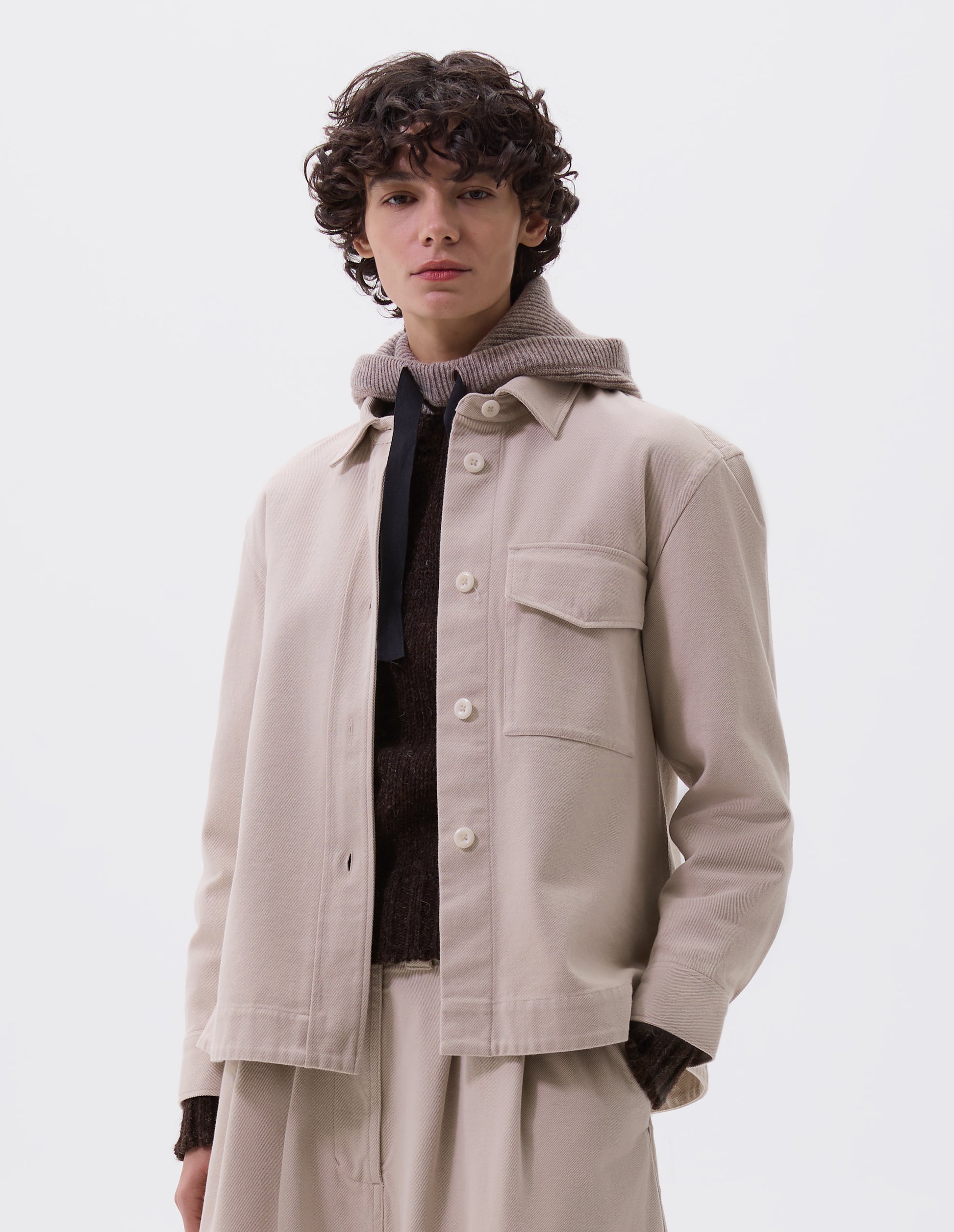 Cropped body image, front view of model wearing beige oversized shirt with stitch through raised placket, corozo button fastening, patch chest pocket and deep stitch hem. Styled with matching skirt, brown jumper and beige hooded scarf.