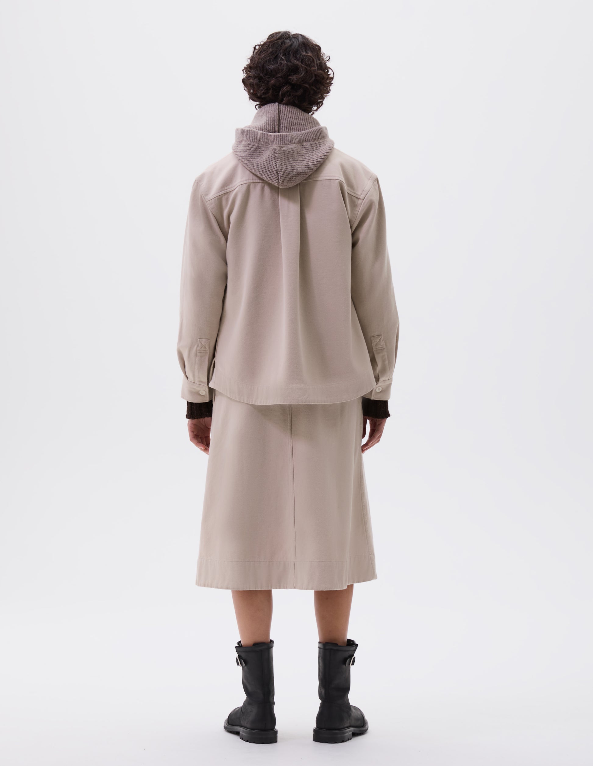 Full body image, back view of model wearing beige oversized shirt with corozo button fastening cuffs, box pleat on back yoke and deep stitch hem. Styled with matching skirt,  beige hood and black leather boots.