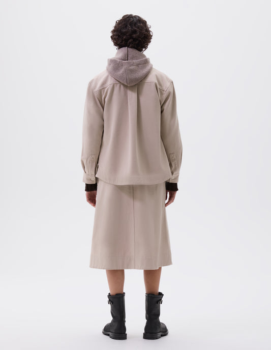 Full body image, back view of model wearing beige oversized shirt with corozo button fastening cuffs, box pleat on back yoke and deep stitch hem. Styled with matching skirt,  beige hood and black leather boots.