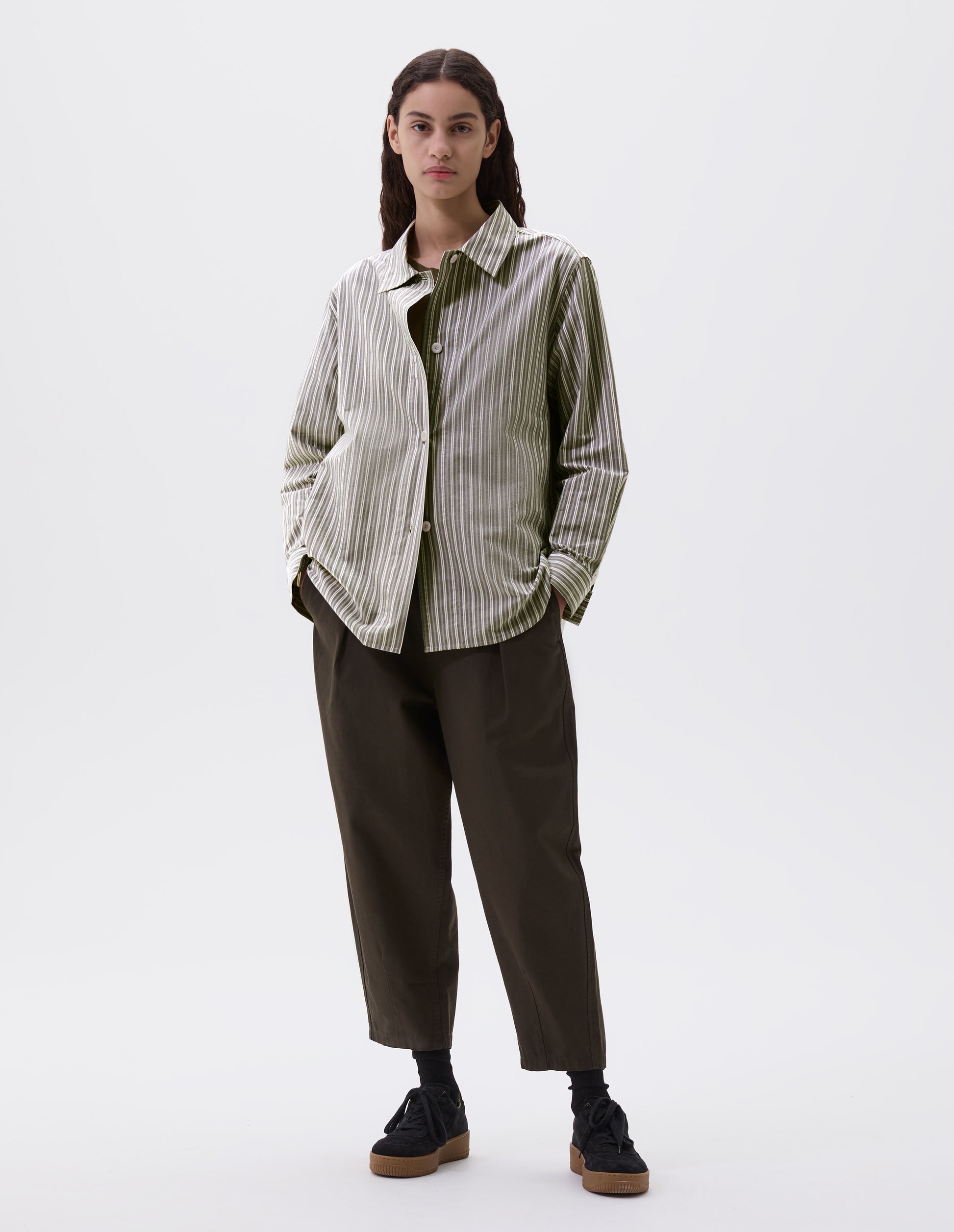 Full body image, front view of model wearing green, grey and off white striped long sleeve shirt. With wide stitch through placket and corozo brace buttons and cuffs. Styled over green t shirt, with green trousers and black nubuck leather trainers.