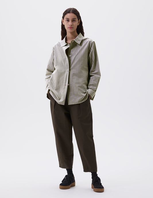 Full body image, front view of model wearing green, grey and off white striped long sleeve shirt. With wide stitch through placket and corozo brace buttons and cuffs. Styled over green t shirt, with green trousers and black nubuck leather trainers.