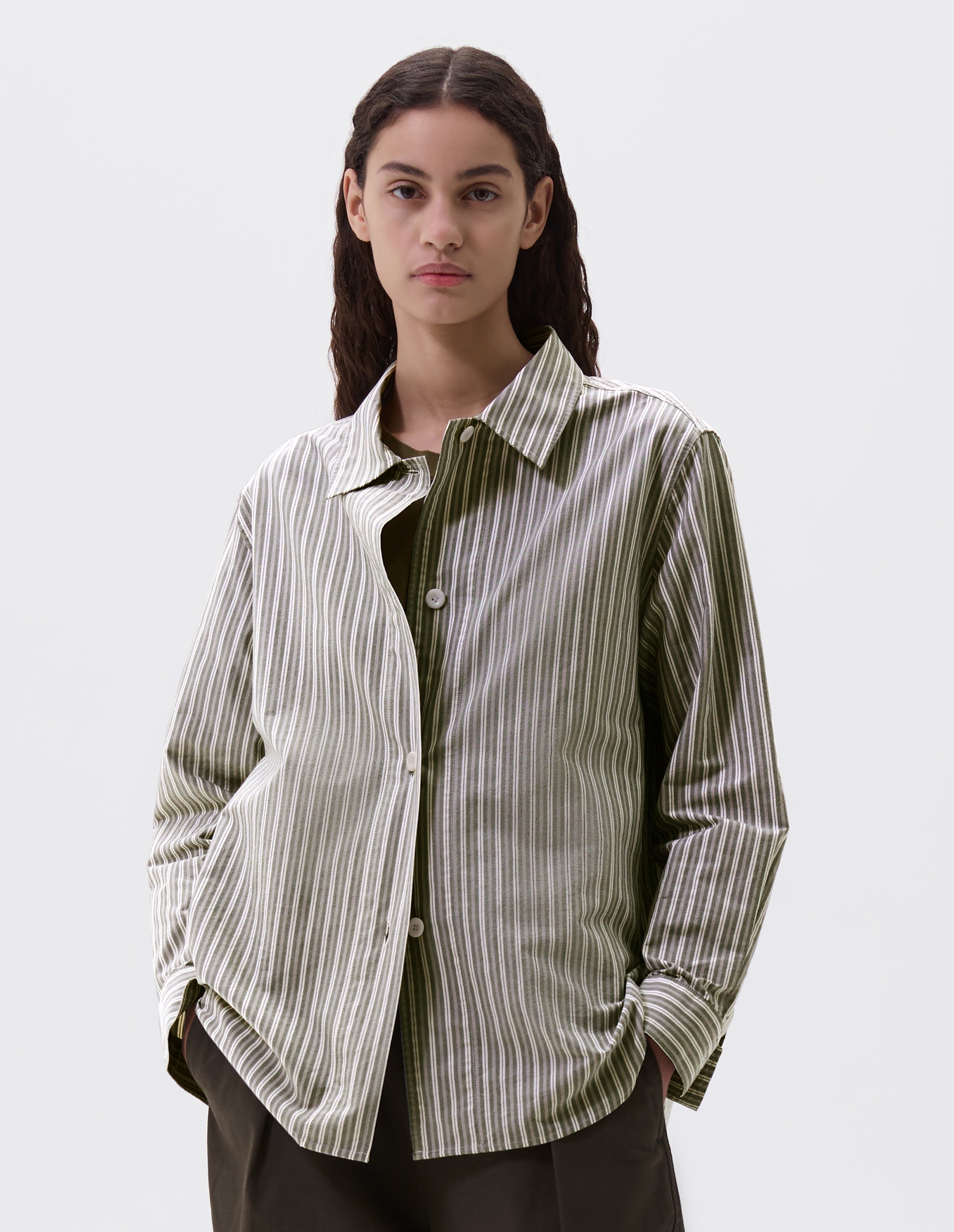 Cropped body image, front view of model wearing green, grey and off white striped long sleeve shirt. With wide stitch through placket and corozo brace buttons and cuffs. Styled over green t shirt, with green trousers.