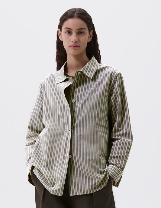 Cropped body image, front view of model wearing green, grey and off white striped long sleeve shirt. With wide stitch through placket and corozo brace buttons and cuffs. Styled over green t shirt, with green trousers.