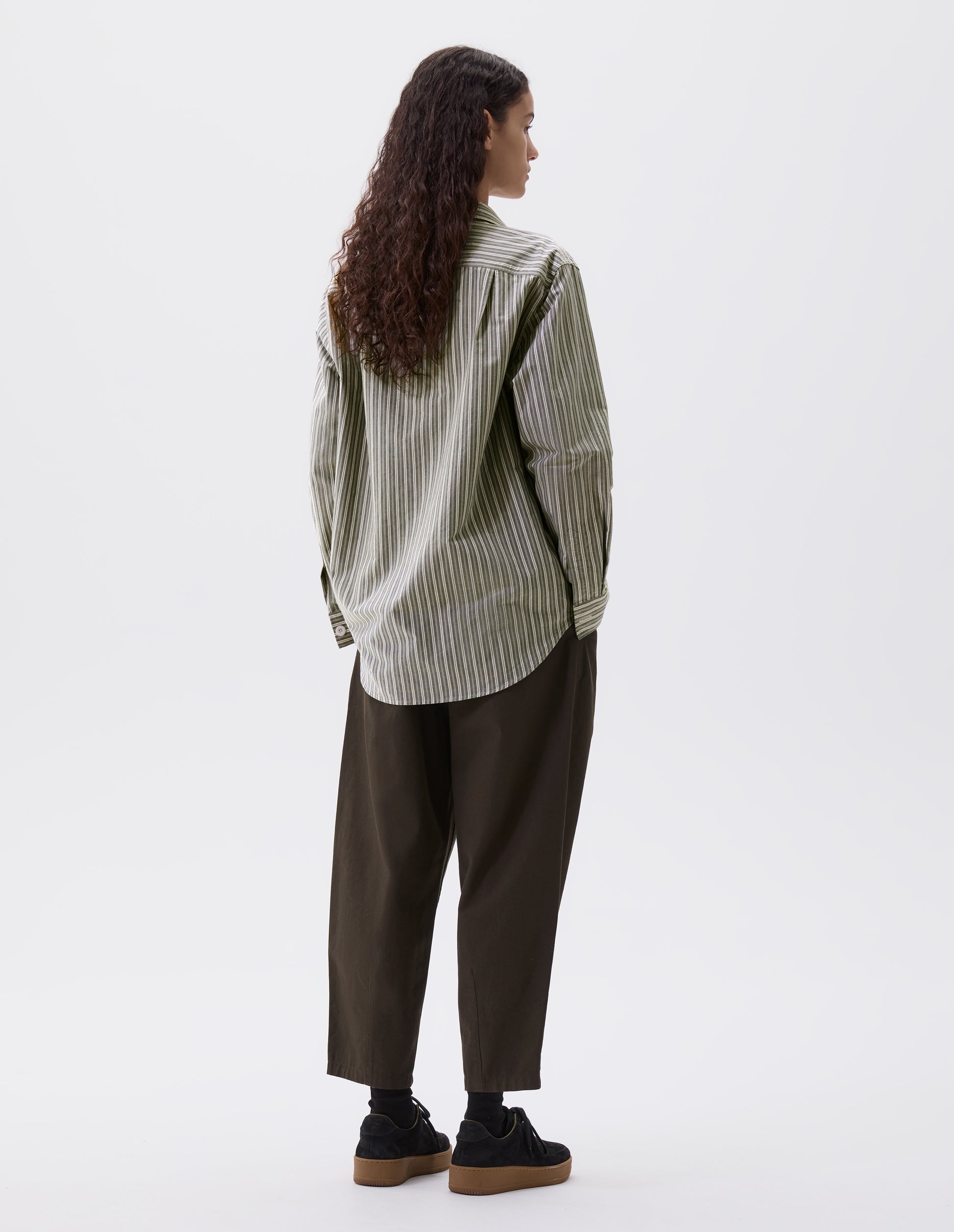 Full body image, back view of model wearing green, grey and off white striped long sleeve shirt. With corozo brace button cuffs and pleated back yoke. Styled with green trousers and black nubuck leather trainers.