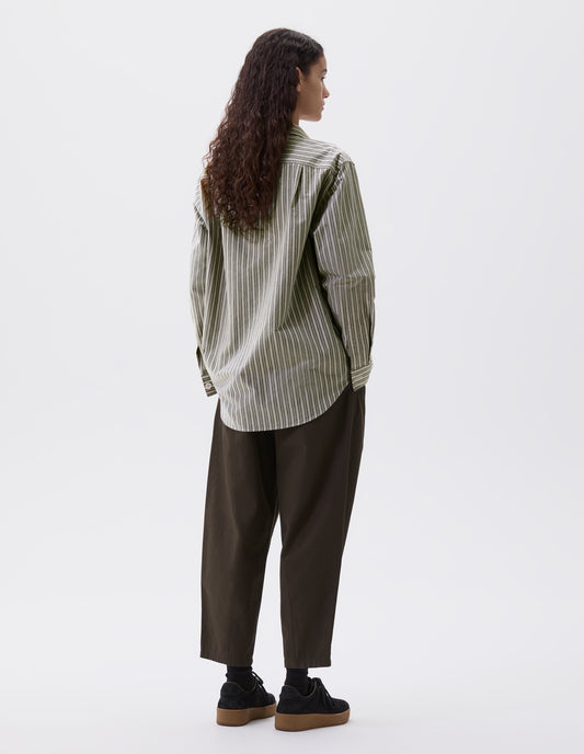 Full body image, back view of model wearing green, grey and off white striped long sleeve shirt. With corozo brace button cuffs and pleated back yoke. Styled with green trousers and black nubuck leather trainers.