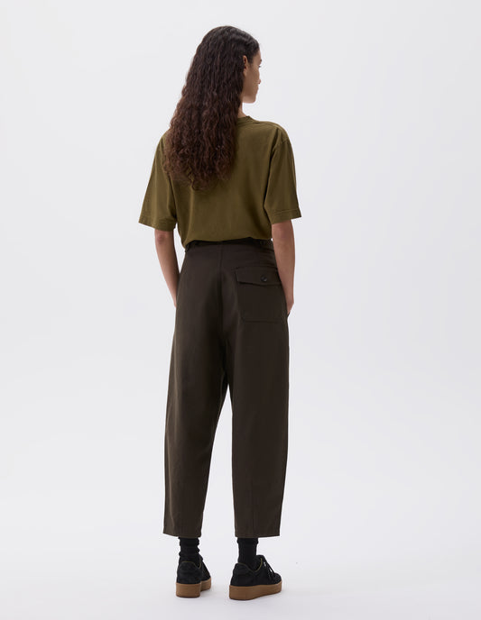 Full body image, back view of model wearing dark green pleated trousers, with button tabs and patch flap button pocket. Styled with olive green t-shirt, black socks and black nubuck trainers. 