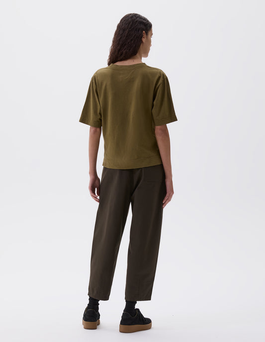Full body image, back view of model wearing green boxy t-shirt. Styled with cropped dark brown trousers, black socks and black nubuck trainers.
