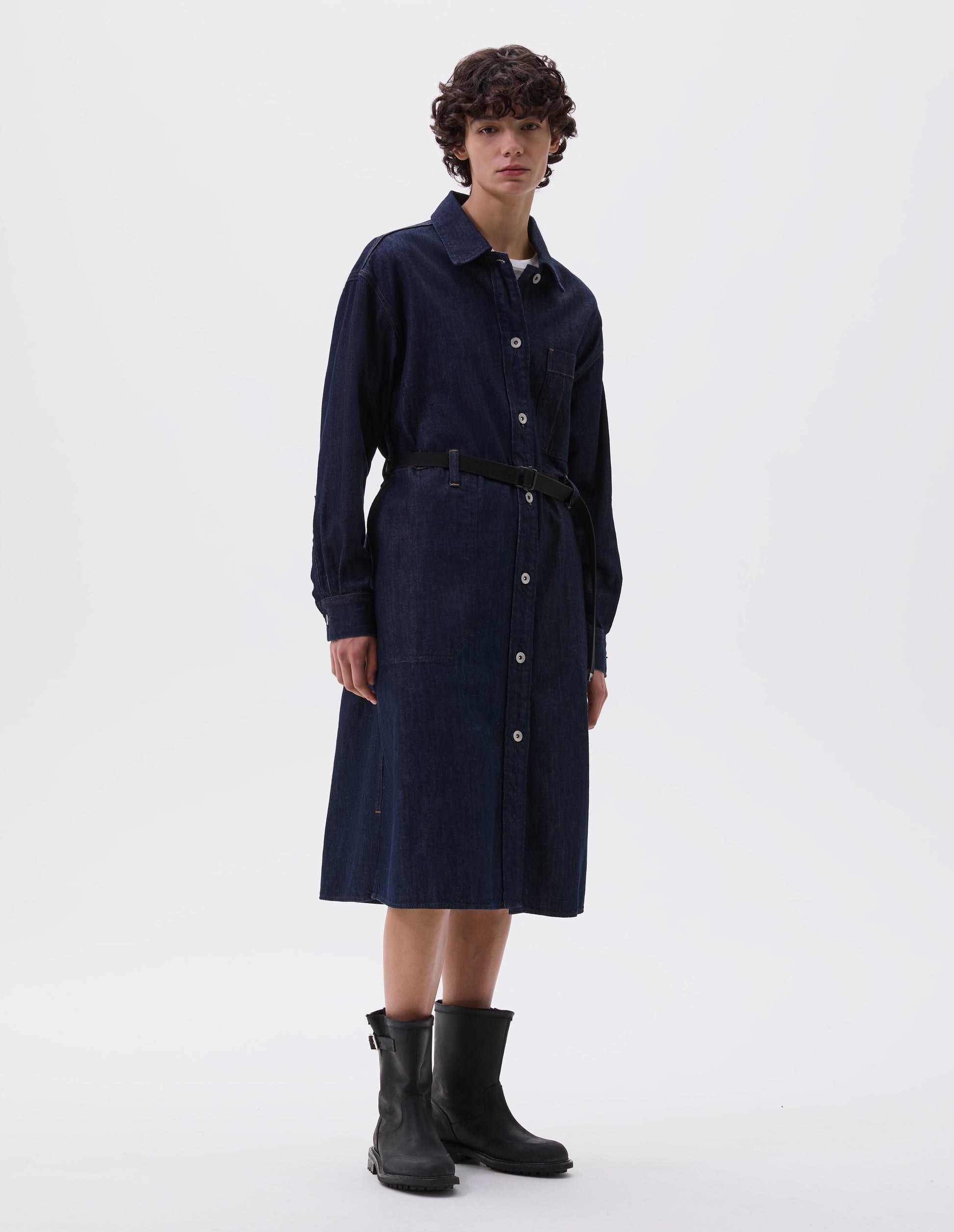 Full body image, front view of model wearing indigo blue denim twill shirt dress with stitch through raised placket, patch pocket with pen pocket detail, slant through side pockets, waist seam detailing with belt loops, side split hem, shirt cuffs and metal button closure. Styled over white t-shirt and with black leather belt and black leather boots.
