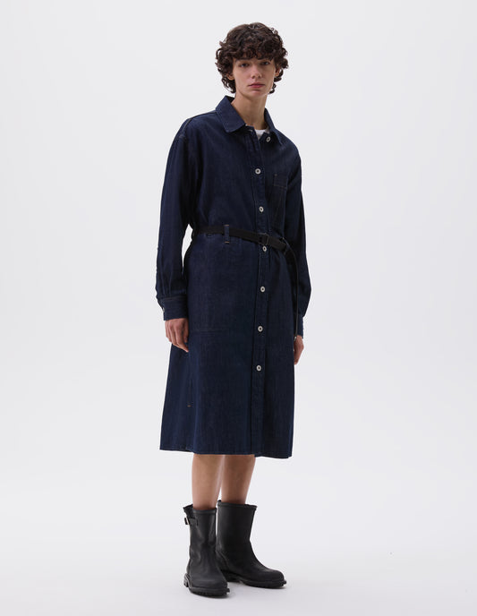 Full body image, front view of model wearing indigo blue denim twill shirt dress with stitch through raised placket, patch pocket with pen pocket detail, slant through side pockets, waist seam detailing with belt loops, side split hem, shirt cuffs and metal button closure. Styled over white t-shirt and with black leather belt and black leather boots.