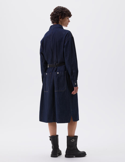 Full body image, back view of model wearing indigo blue denim twill shirt dress with waist seam detailing with belt loops, side split hem, shirt cuffs with metal buttons and patch pockets on back skirt with metal buttons. Styled with black leather belt and black leather boots.