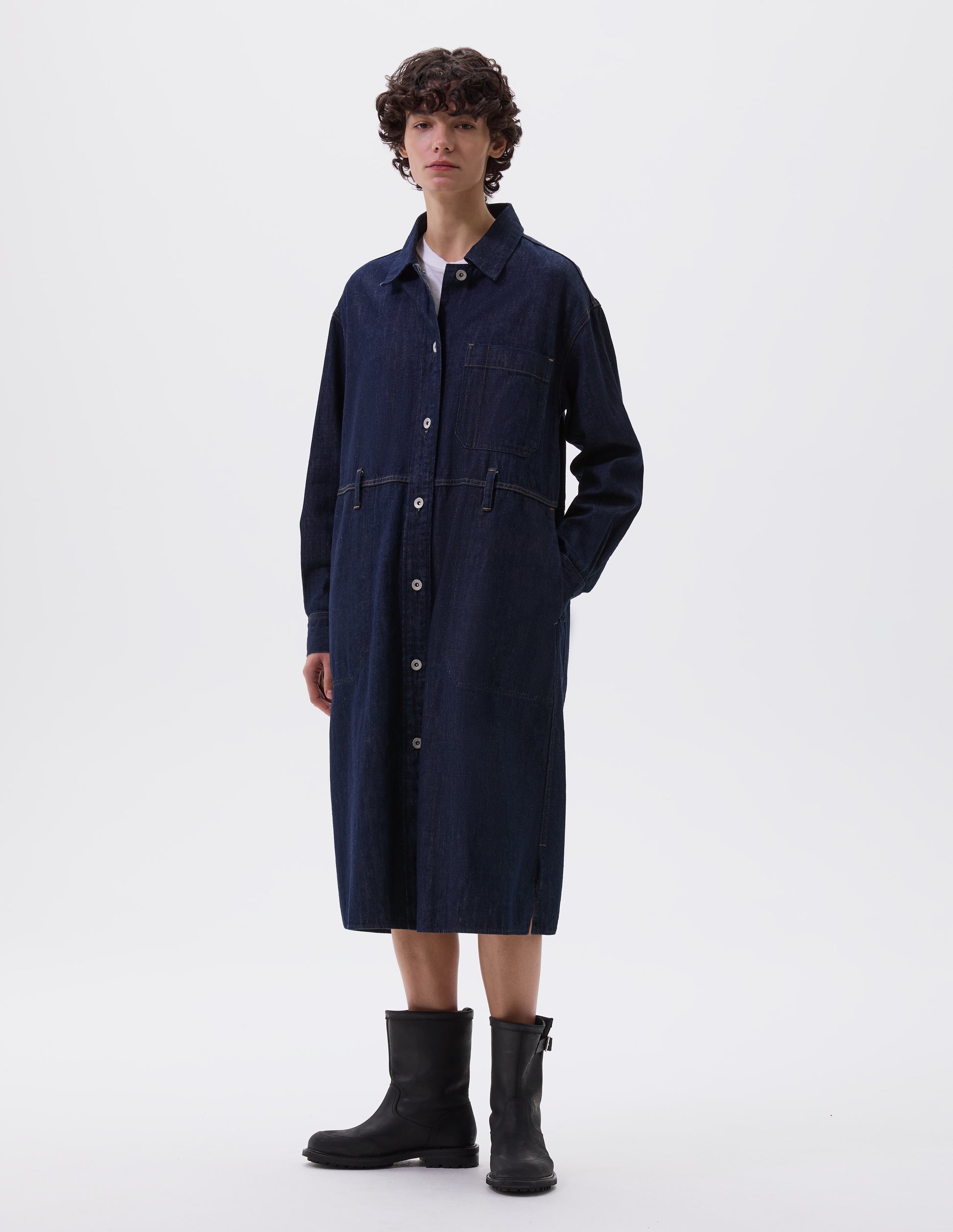 Full body image, front view of model wearing indigo blue denim twill shirt dress with stitch through raised placket, patch pocket with pen pocket detail, slant through side pockets, waist seam detailing with belt loops, side split hem, shirt cuffs and metal button closure. Styled over white t-shirt and with black leather boots.
