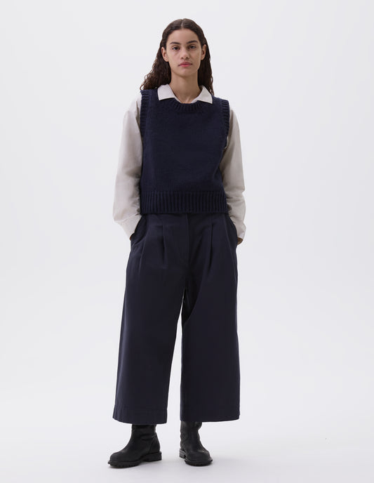 Full body image, front view of model wearing navy blue sleeveless knitted vest with contrasting neckline, hem and deep armholes. Styled over a light grey shirt, navy blue pleated trousers and black leather boots. 