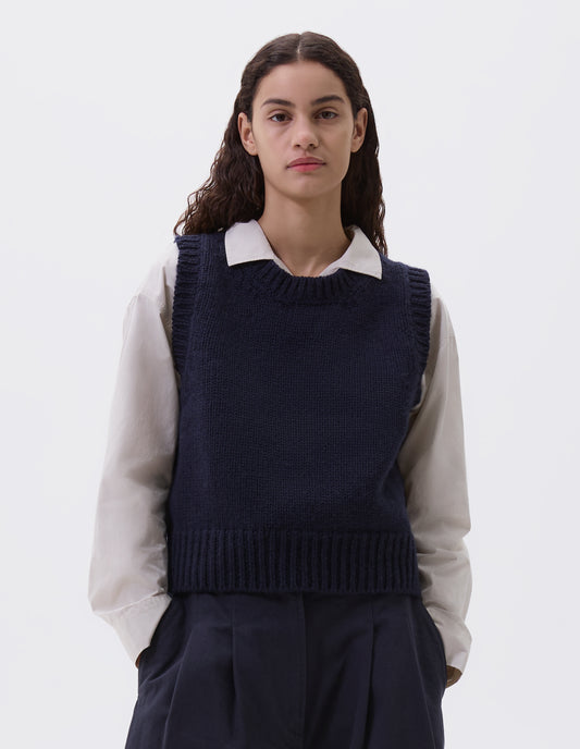 Cropped body image, front view of model wearing navy blue sleeveless knitted vest with contrasting neckline, hem and deep armholes. Styled over a light grey shirt with navy blue pleated trousers.