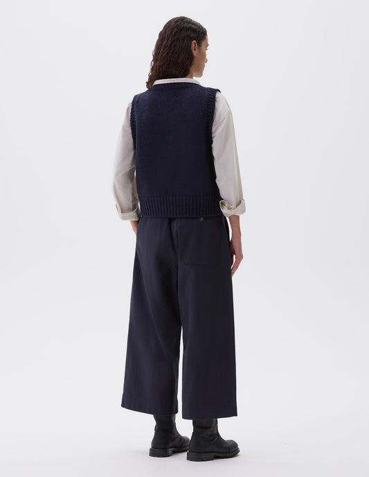 Full body image, back view of model wearing dark blue double pleated culottes, with half elasticated waist, belt loops and press stud patch back pocket. Styled with dark blue slipover over light grey shirt and black leather boots.