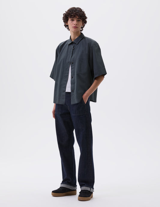 Full body image, front view of model wearing greenish blue shirt with elbow length sleeves, patch chest pocket and vent split hem. Styled over white t-shirt, with indigo blue jeans and black suede trainers. 