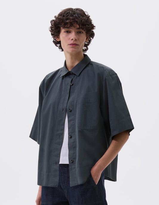 Cropped body image, front view of model wearing greenish blue shirt with elbow length sleeves, patch chest pocket and vent split hem. Styled over white t-shirt, with indigo blue jeans.