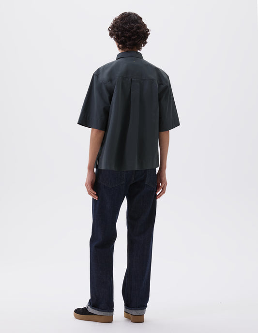 Full body image, back view of model wearing greenish blue shirt with elbow length sleeves, vent split hem and hanger loop on back yoke. Styled with indigo blue jeans and black suede trainers. 