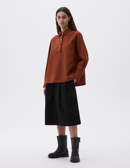 Full body image, front view of model wearing brownish red shirt with half placket, large chest pocket and corozo button cuffs. Styled with black denim skirt and black leather boots.