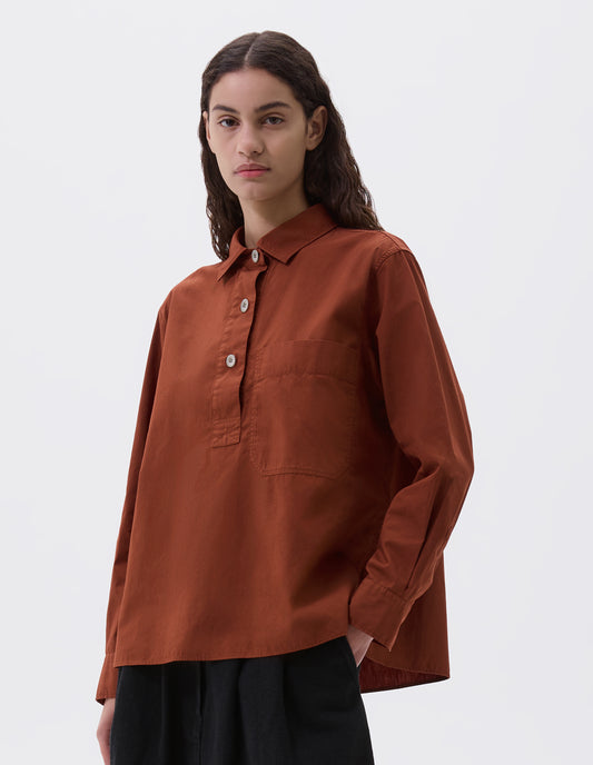 Cropped body image, front view of model wearing brownish red shirt with half placket, large chest pocket and corozo button cuffs. Styled with black denim skirt.