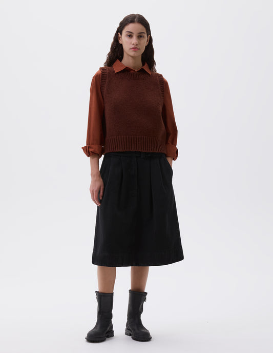 Full body image, front view of model wearing red brown sleeveless knitted vest with contrasting neckline, hem and deep armholes. Styled with orange long sleeve shirt, black pleated skirt and black leather boots.
