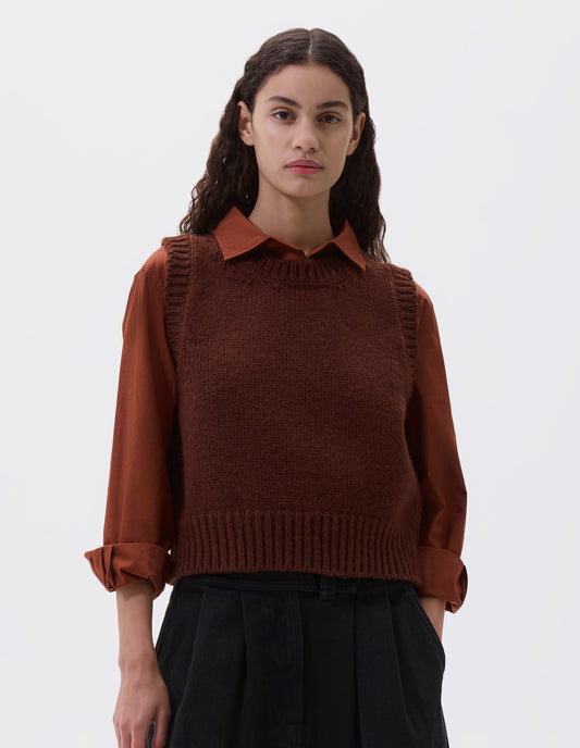 Cropped body image, front view of model wearing red brown sleeveless knitted vest with contrasting neckline, hem and deep armholes. Styled with orange long sleeve shirt and black pleated skirt.