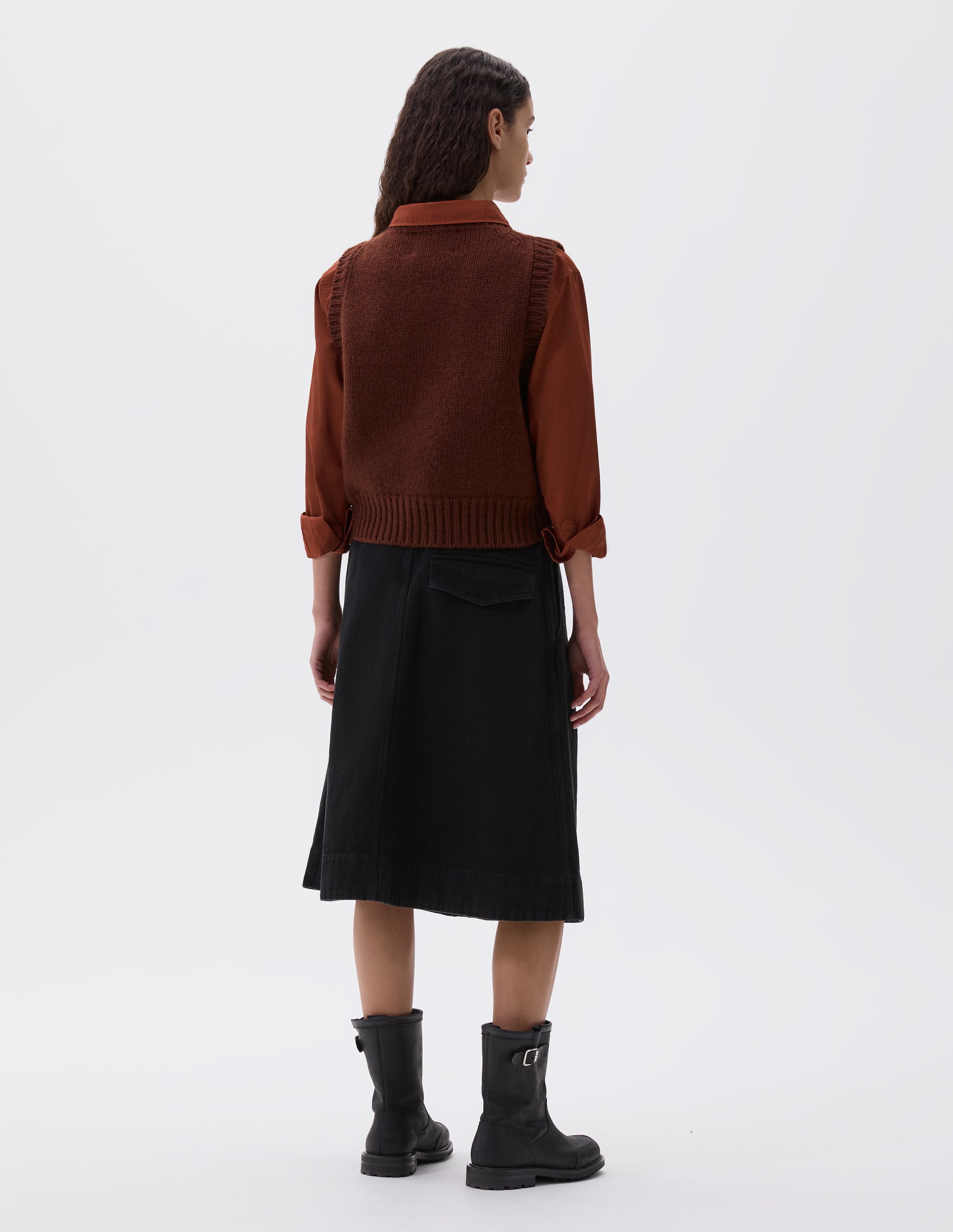 Full body image, back view of model wearing red brown sleeveless knitted vest with contrasting neckline, hem and deep armholes. Styled with orange long sleeve shirt, black pleated skirt and black leather boots.