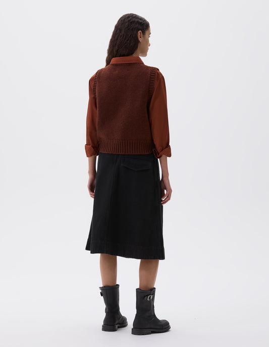 Full body image, back view of model wearing red brown sleeveless knitted vest with contrasting neckline, hem and deep armholes. Styled with orange long sleeve shirt, black pleated skirt and black leather boots.