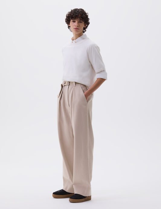 Full body image, front view of model wearing beige wide leg trousers with button fastening waistband, slant pockets and removable webbing belt with metal slide adjuster. Styled with white shirt, layered underneath a white t-shirt and black suede trainers.