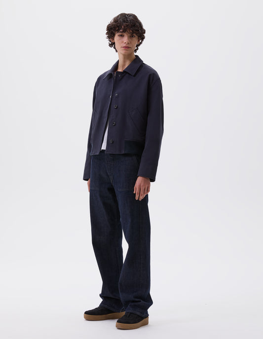 Full body image, front view of model wearing dark navy collared jacket with press stud closure, slant deep welt pockets and ribbed hem. Styled with white t-shirt and indigo blue denim jeans and black nubuck trainers. 