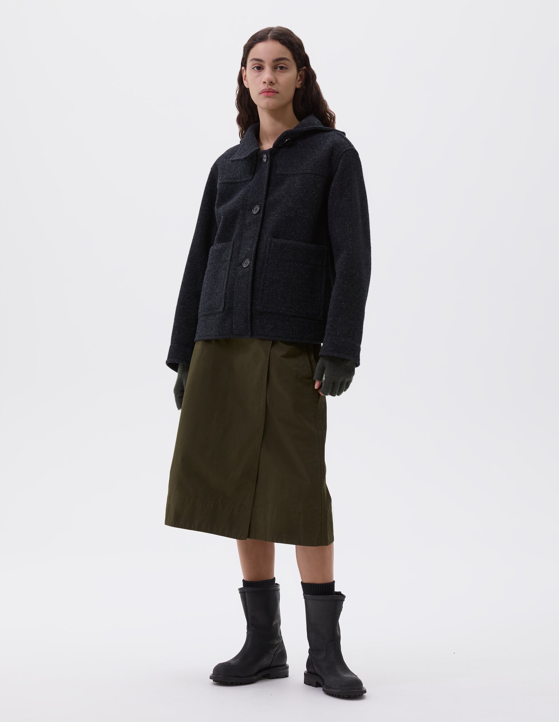 Full body image, front view of model wearing charcoal grey short hooded duffle coat, with button fastening storm tab, patch pockets and front yoke detailing. Styled with brown fingerless gloves, olive green skirt, black socks and black leather boots.