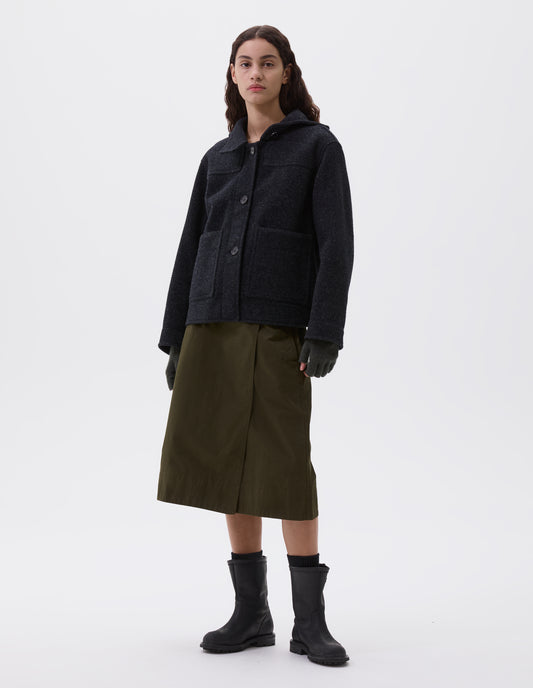 Full body image, front view of model wearing charcoal grey short hooded duffle coat, with button fastening storm tab, patch pockets and front yoke detailing. Styled with brown fingerless gloves, olive green skirt, black socks and black leather boots.