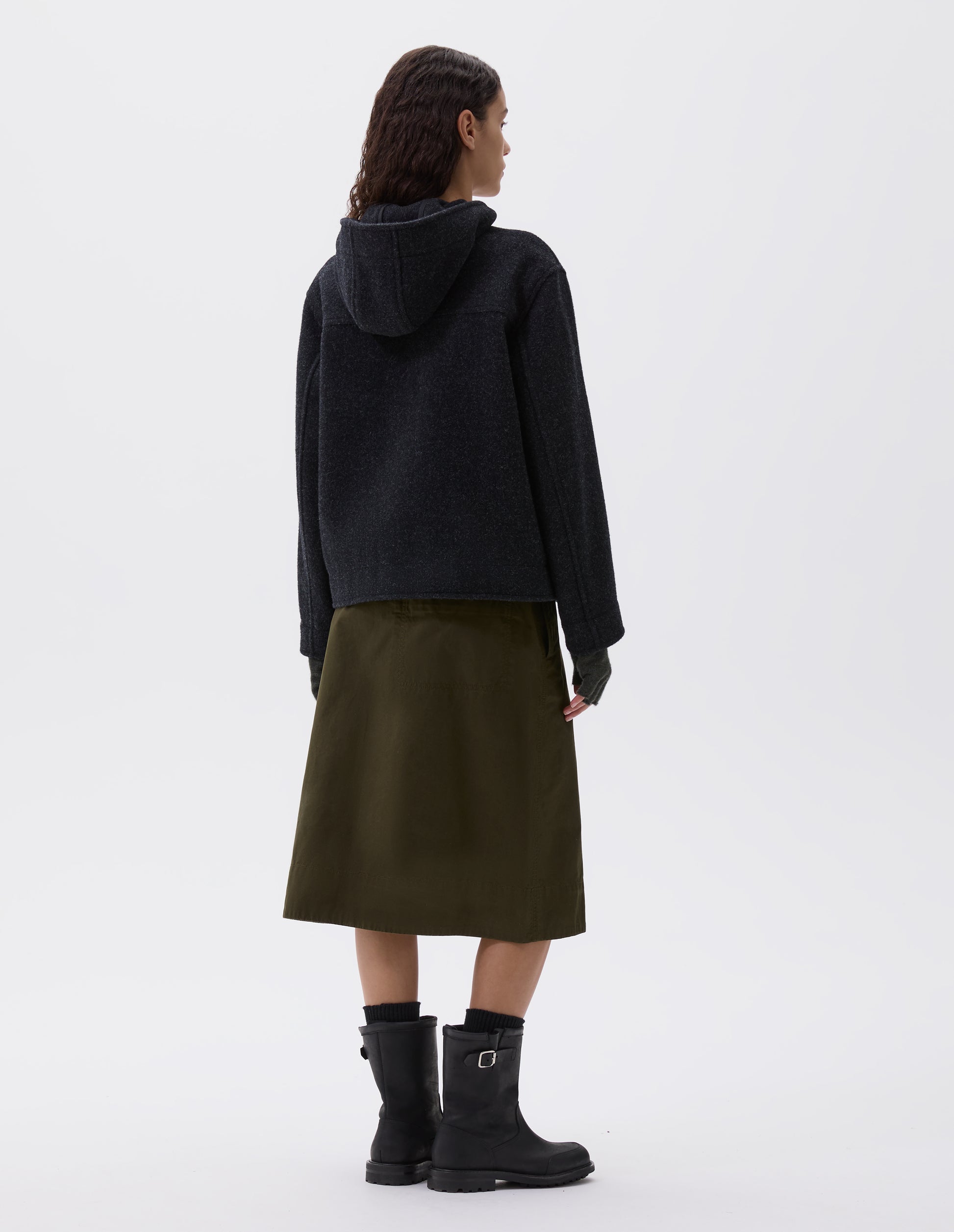 Full body image, back view of model wearing charcoal grey short hooded duffle coat, with back yoke detailing. Styled with brown fingerless gloves, olive green skirt, black socks and black leather boots.
