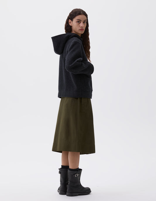 Full body image, side view of model wearing charcoal grey short hooded duffle coat, with patch pockets. Styled with olive green skirt, black socks and black leather boots.
