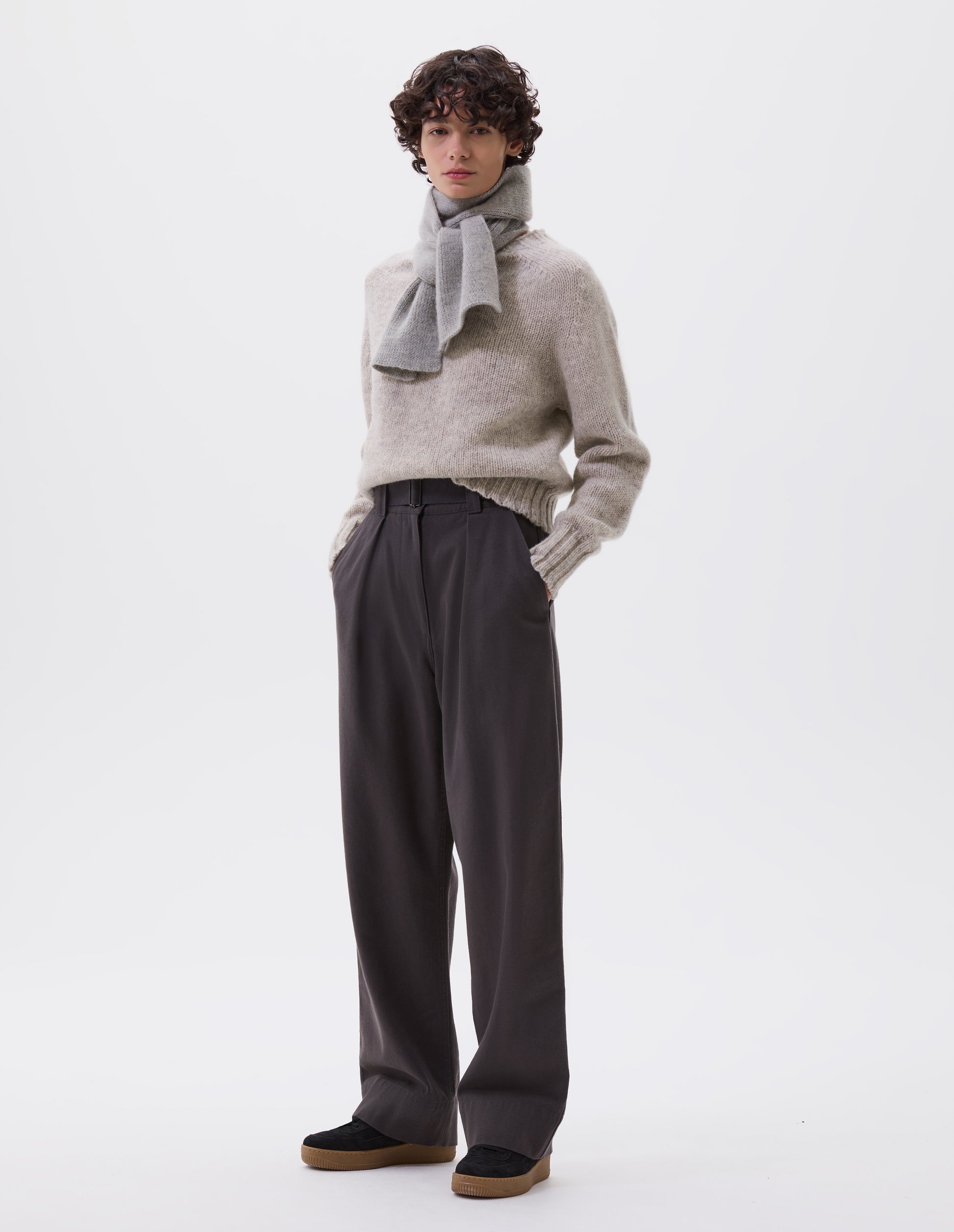 Full body image, front view of model wearing charcoal grey wide leg trousers with button fastening waistband, slant pockets and removable webbing belt with metal slide adjuster. Styled with beige jumper, grey scarf and black suede trainers.