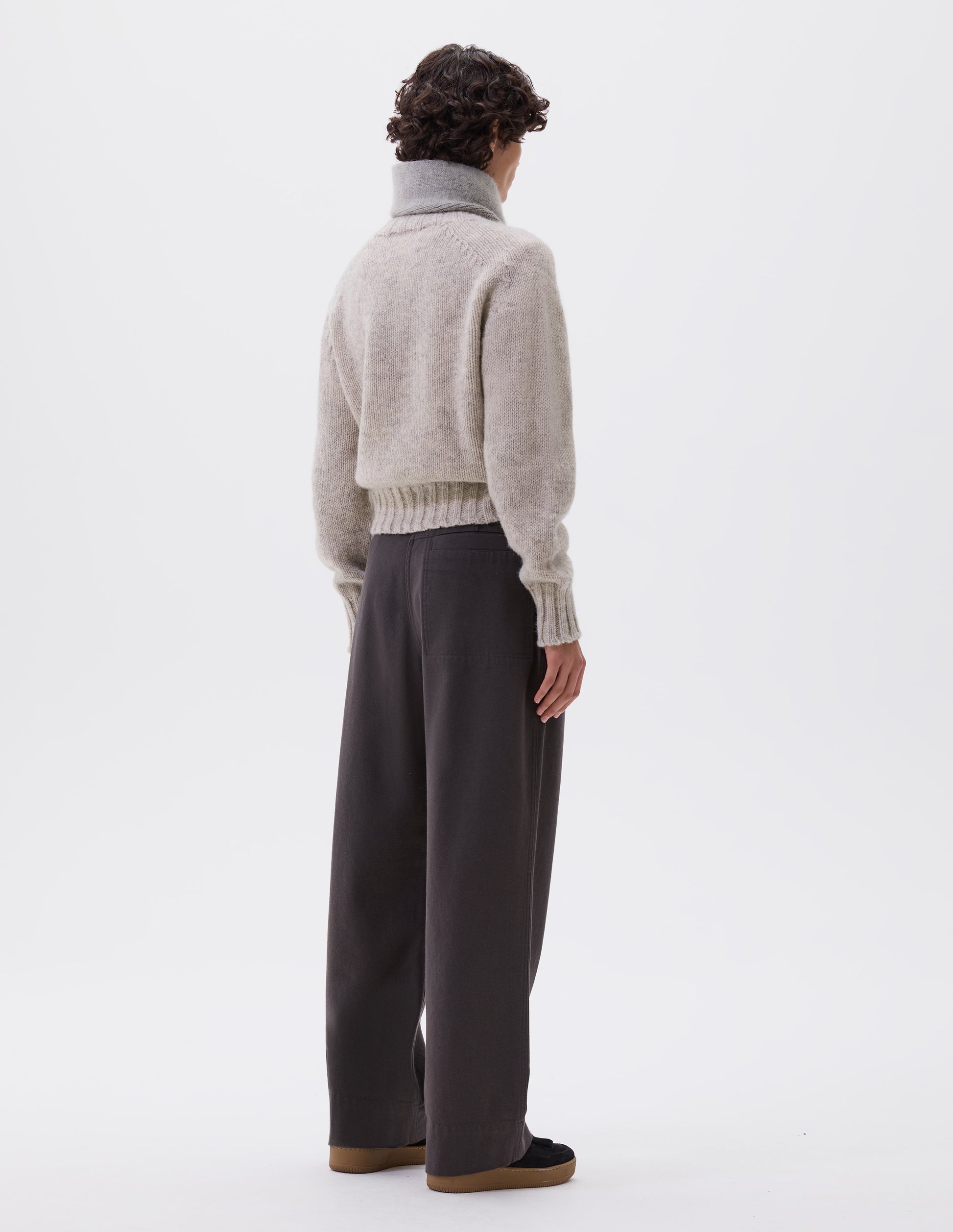 Full body image, back view of model wearing charcoal grey wide leg trousers with stitch through back welt pocket, belt loops and removable webbing belt with metal slide adjuster. Styled with beige jumper, grey scarf and black suede trainers.