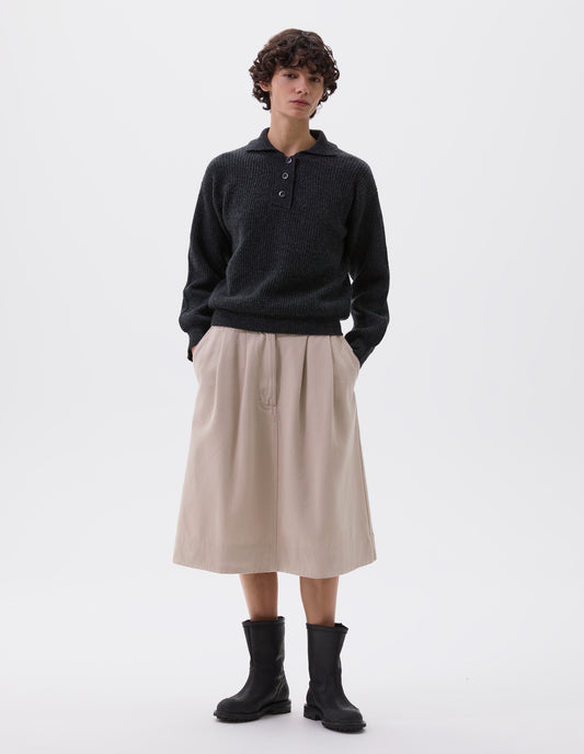 Full body image, front view of model wearing dark grey knitted polo with corozo brace buttons and contrast rib on collar, placket and cuffs. Styled with beige midi skirt with side pockets and black leather biker boots.