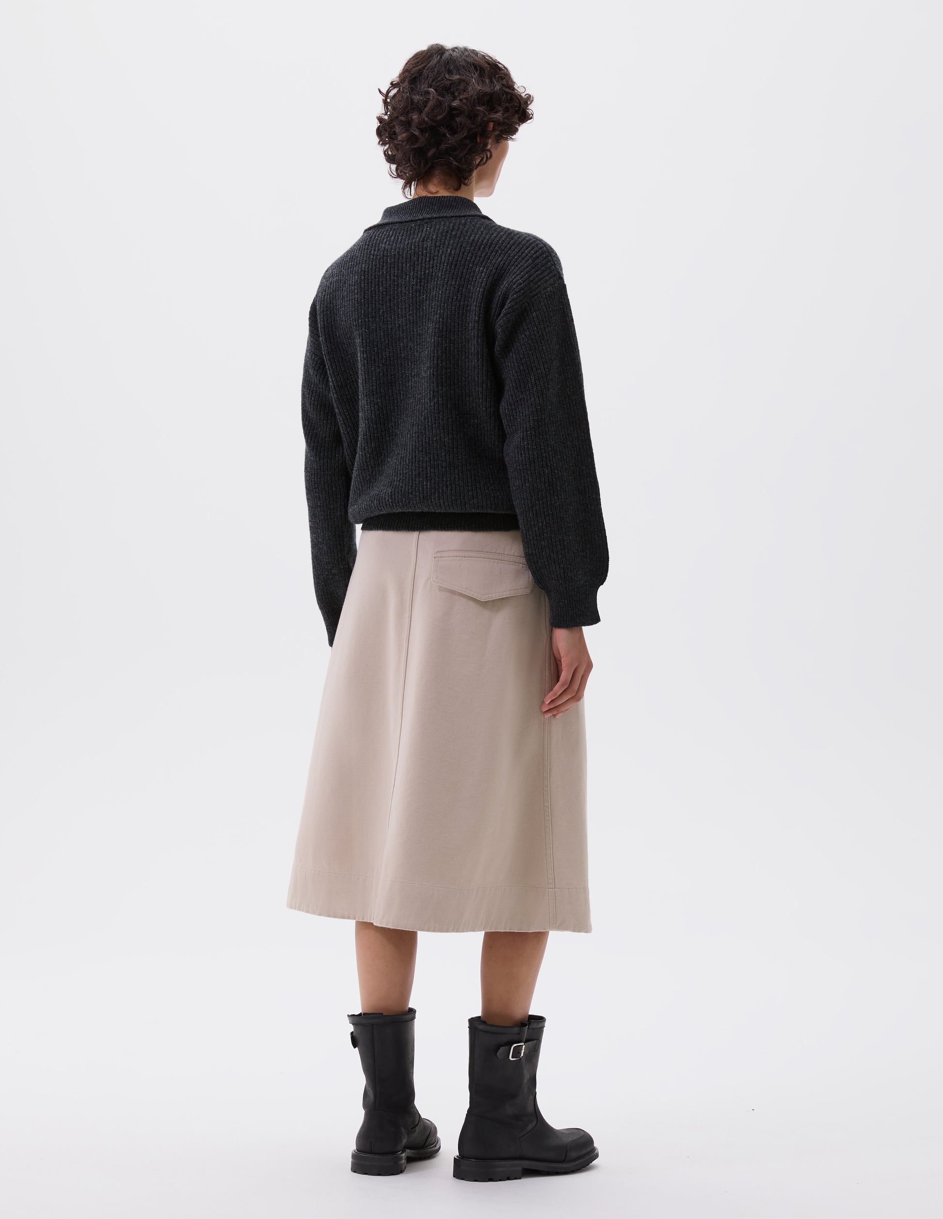 Full body image, back view of model wearing beige A-line skirt with deep stitch hem and back flap pocket. Styled with grey knitted polo and black leather biker boots.