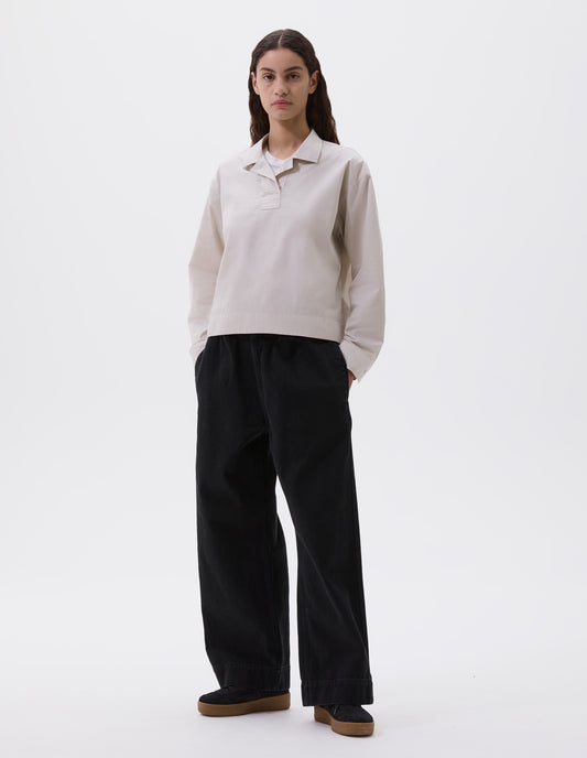 Full body image, front view of model wearing beige cropped open neck pull-on shirt with box stitch detailing on placket. Styled over white t-shirt and with black trousers and black trainers.