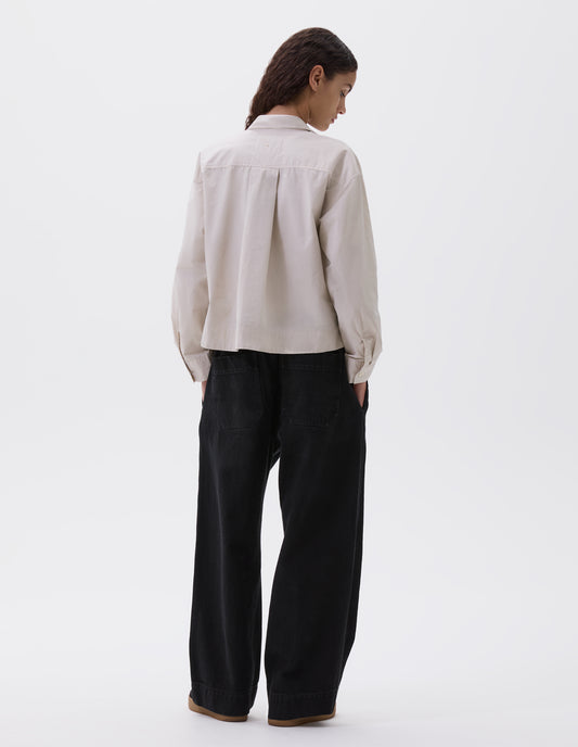 Full body image, back view of model wearing beige cropped shirt with wide box pleat on back yoke and corozo brace buttons on cuffs. Styled with black trousers and black trainers.