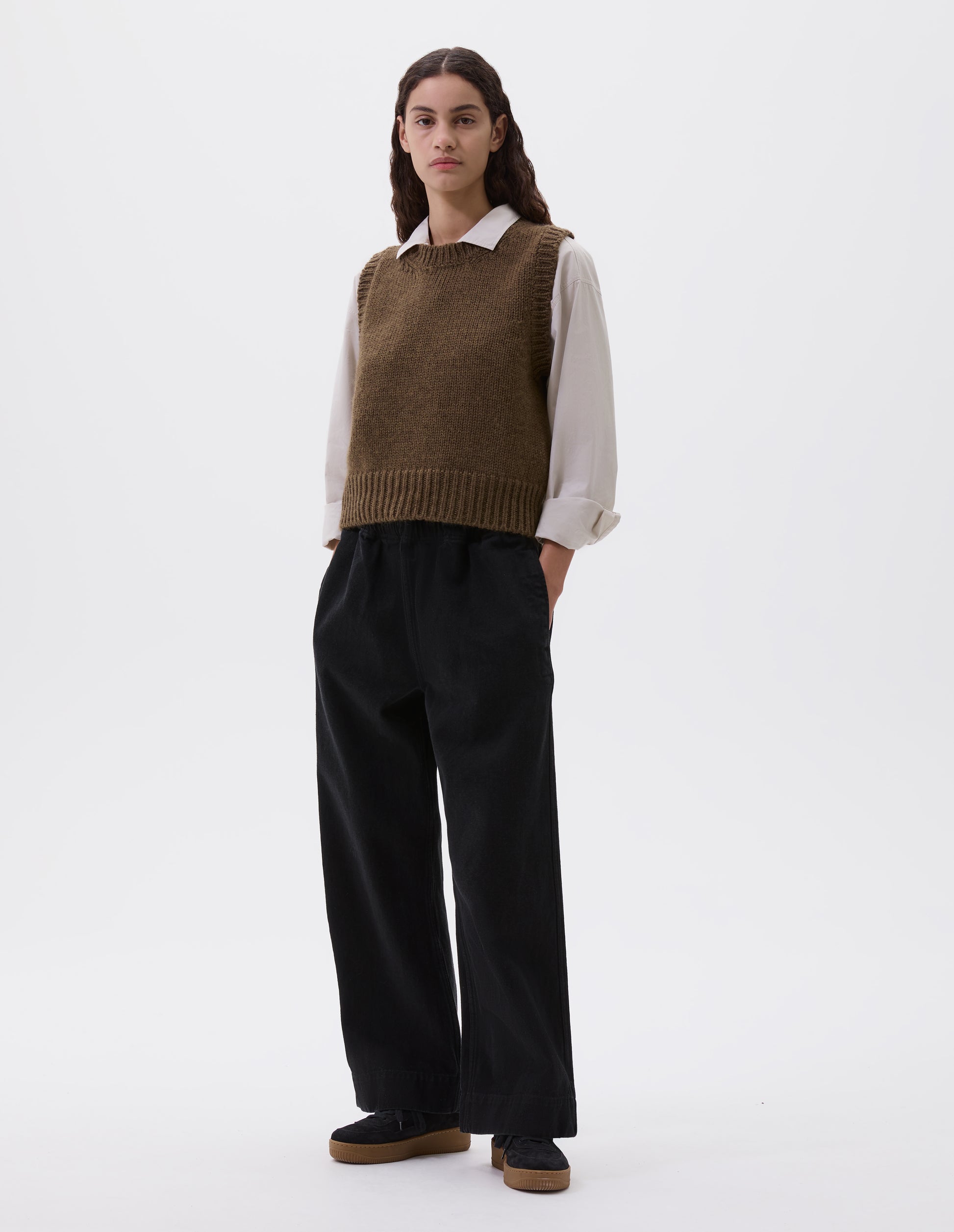 Full body image, front view of model wearing beige green sleeveless knitted vest with contrasting neckline, hem and deep armholes. Styled over a light grey shirt, black wide leg trousers and black nubuck leather trainers.