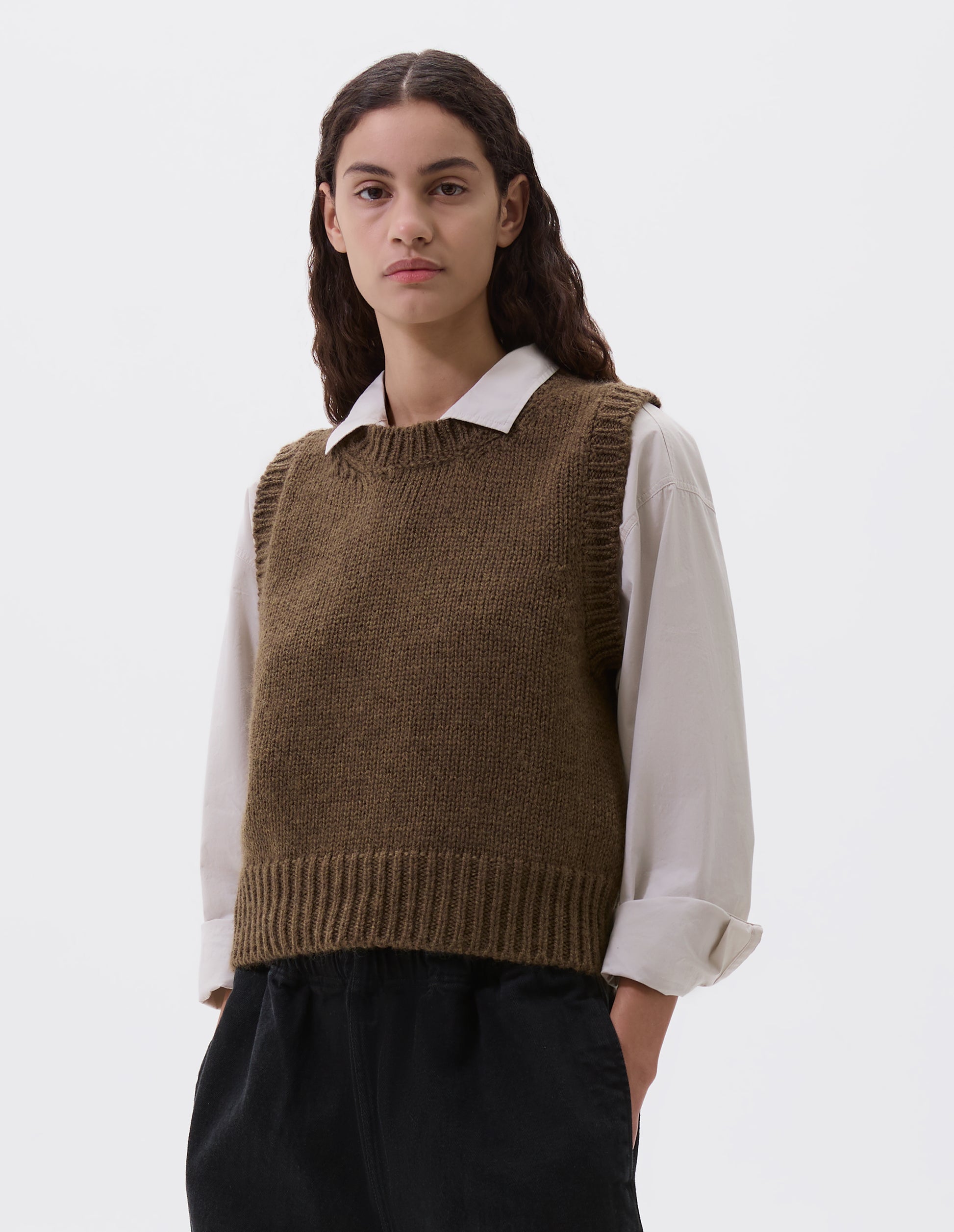 Cropped body image, front view of model wearing beige green sleeveless knitted vest with contrasting neckline, hem and deep armholes. Styled over a light grey shirt and black wide leg trousers.
