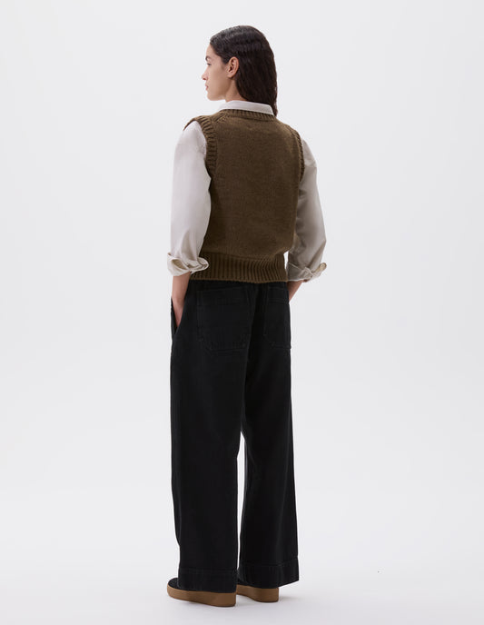 Full body image, back view of model wearing beige green sleeveless knitted vest with contrasting neckline, hem and deep armholes. Styled over a light grey shirt, black wide leg trousers and black nubuck leather trainers.