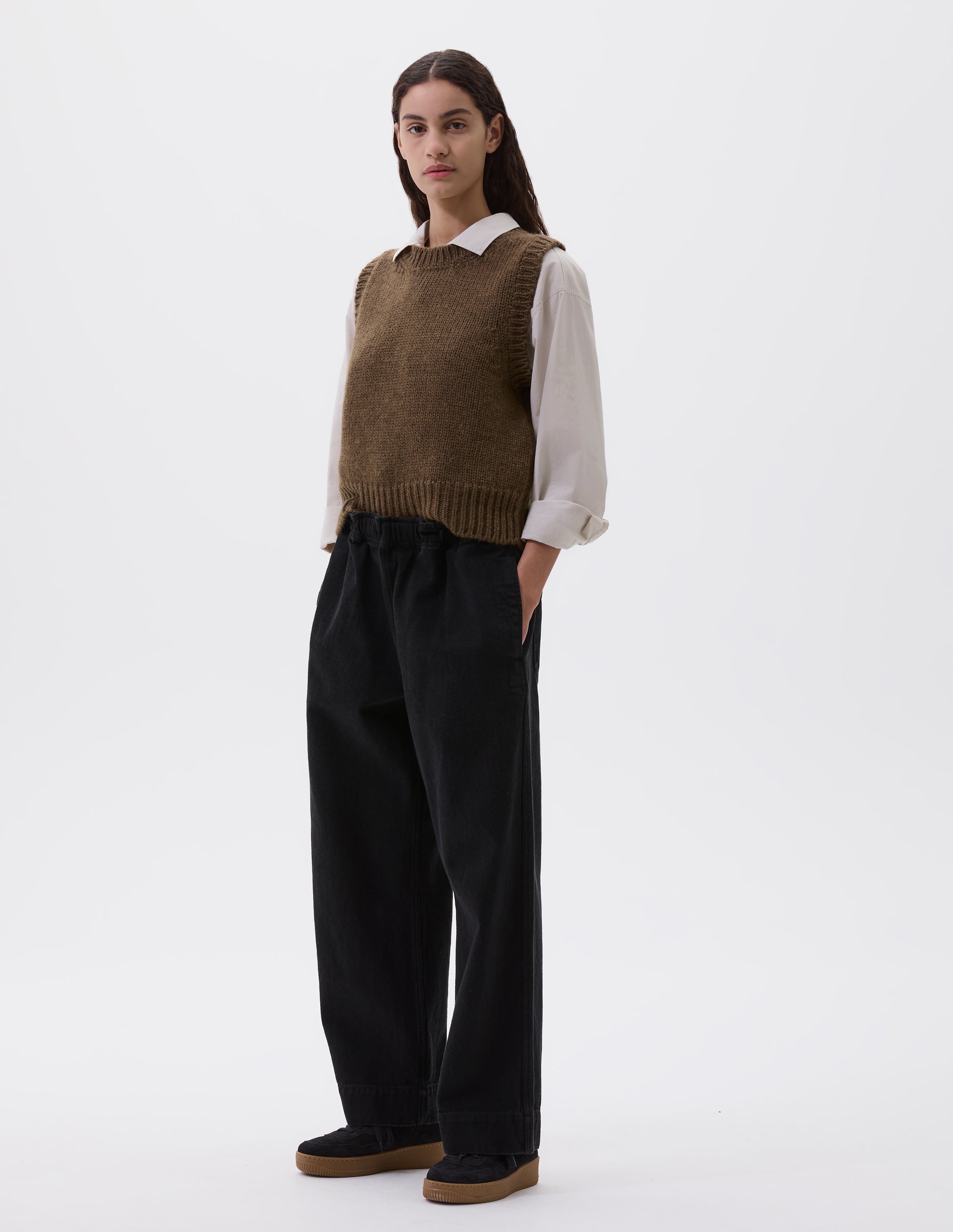 Full body image, front view of model wearing black relaxed wide leg joggers with elasticated waistband and side seam pockets. Styled with light brown slipover, beige shirt and black nubuck trainers.