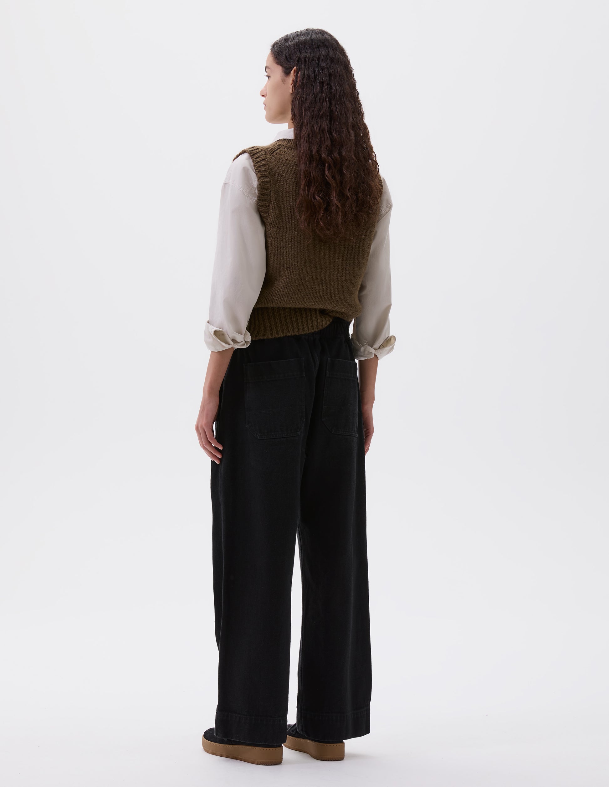 Full body image, back view of model wearing black relaxed wide leg joggers with elasticated waistband and patch back pockets. Styled with light brown slipover, beige shirt and black nubuck trainers.