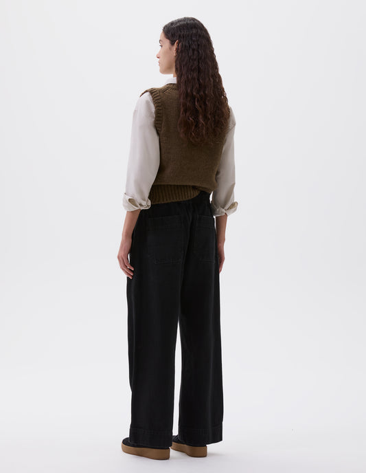 Full body image, back view of model wearing black relaxed wide leg joggers with elasticated waistband and patch back pockets. Styled with light brown slipover, beige shirt and black nubuck trainers.