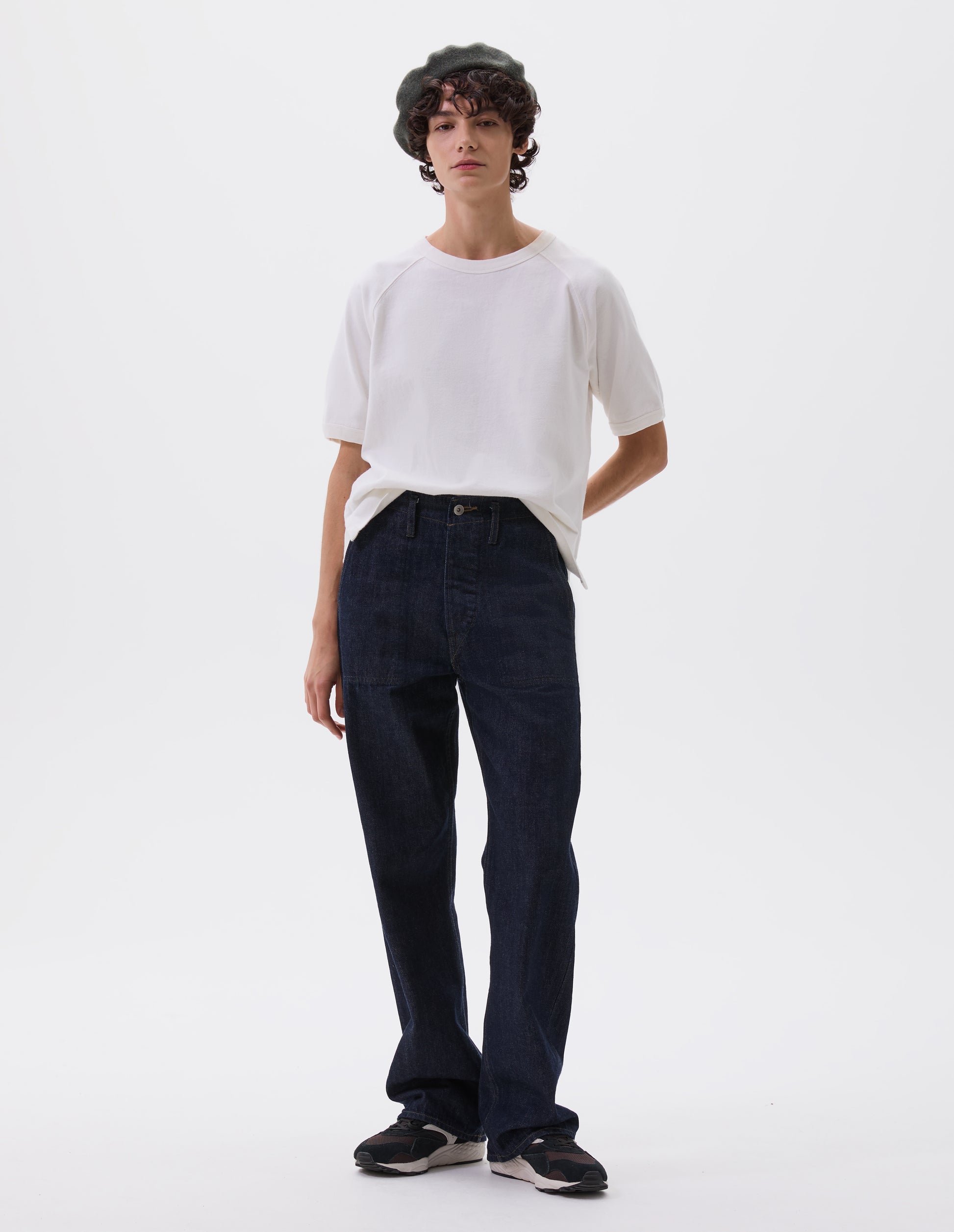 Full body image, front view of model wearing indigo blue denim twill jeans, with metal button fly, stitch-through waistband with belt loops, and stitch through pockets. Styled with white t-shirt and black and brown trainers.