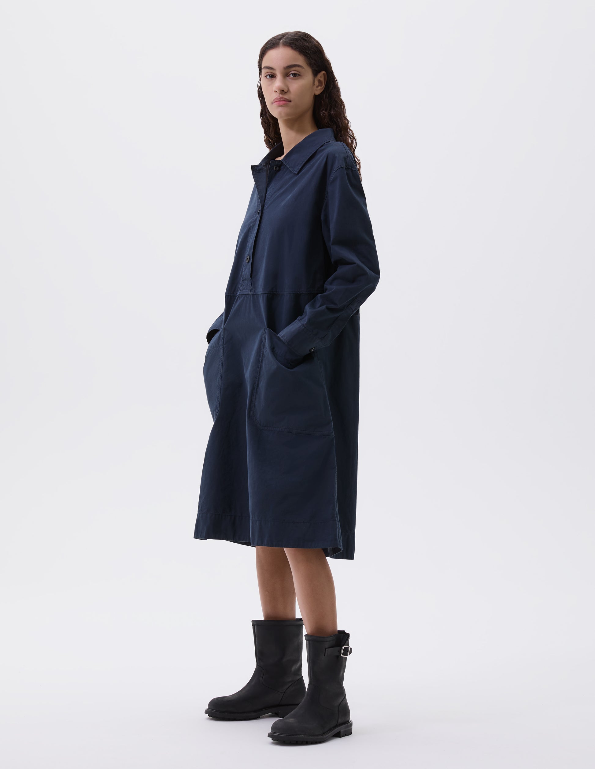 Full body image, front view of model wearing dark blue long sleeve shirt dress with box stitch detailing, large apron patch pockets, deep stitch hem and waist seam detail. Styled with black leather biker boots.